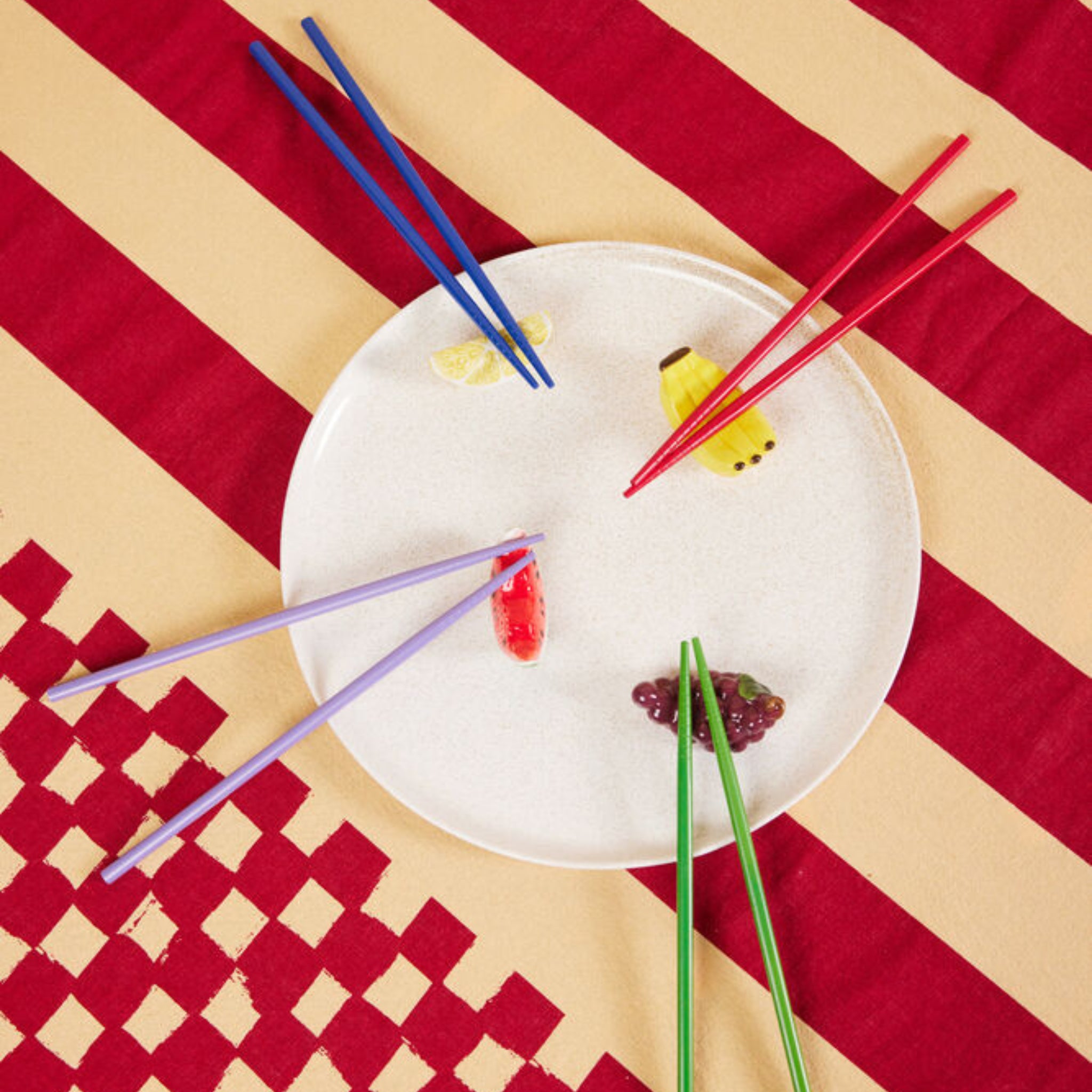 Een wit bord op een rood en geel gestreept tafelkleed met Byon Yaki Chopsticks 4 st. van Byon die voedsel in de vorm van citroen, maïs, watermeloen en druiven vasthouden - een ideaal cadeau voor elke gelegenheid.