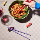 Een hand gebruikt Byon Yaki Chopsticks 4 st. om noedels uit een groene kom te tillen op een geruit tafelkleed. Een wijnglas, een schijfje citroen, een schijfje grapefruit en decoratieve paarse eetstokjes van Byon zorgen voor een uniek cadeau.