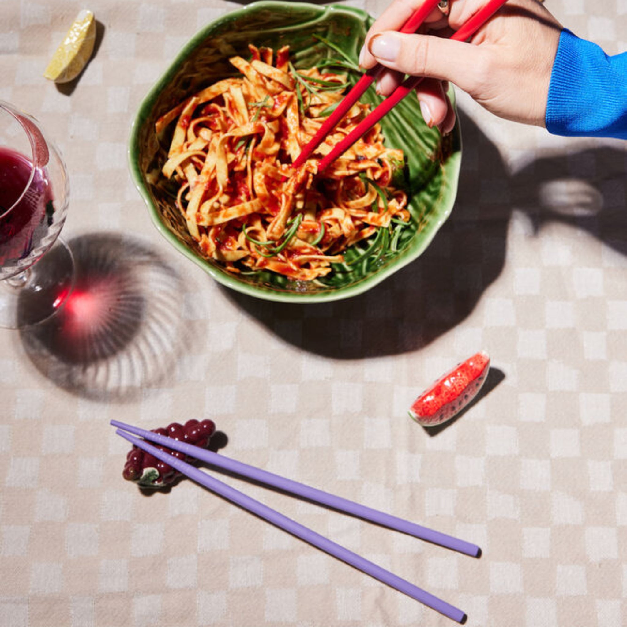 Een hand gebruikt Byon Yaki Chopsticks 4 st. om noedels uit een groene kom te tillen op een geruit tafelkleed. Een wijnglas, een schijfje citroen, een schijfje grapefruit en decoratieve paarse eetstokjes van Byon zorgen voor een uniek cadeau.