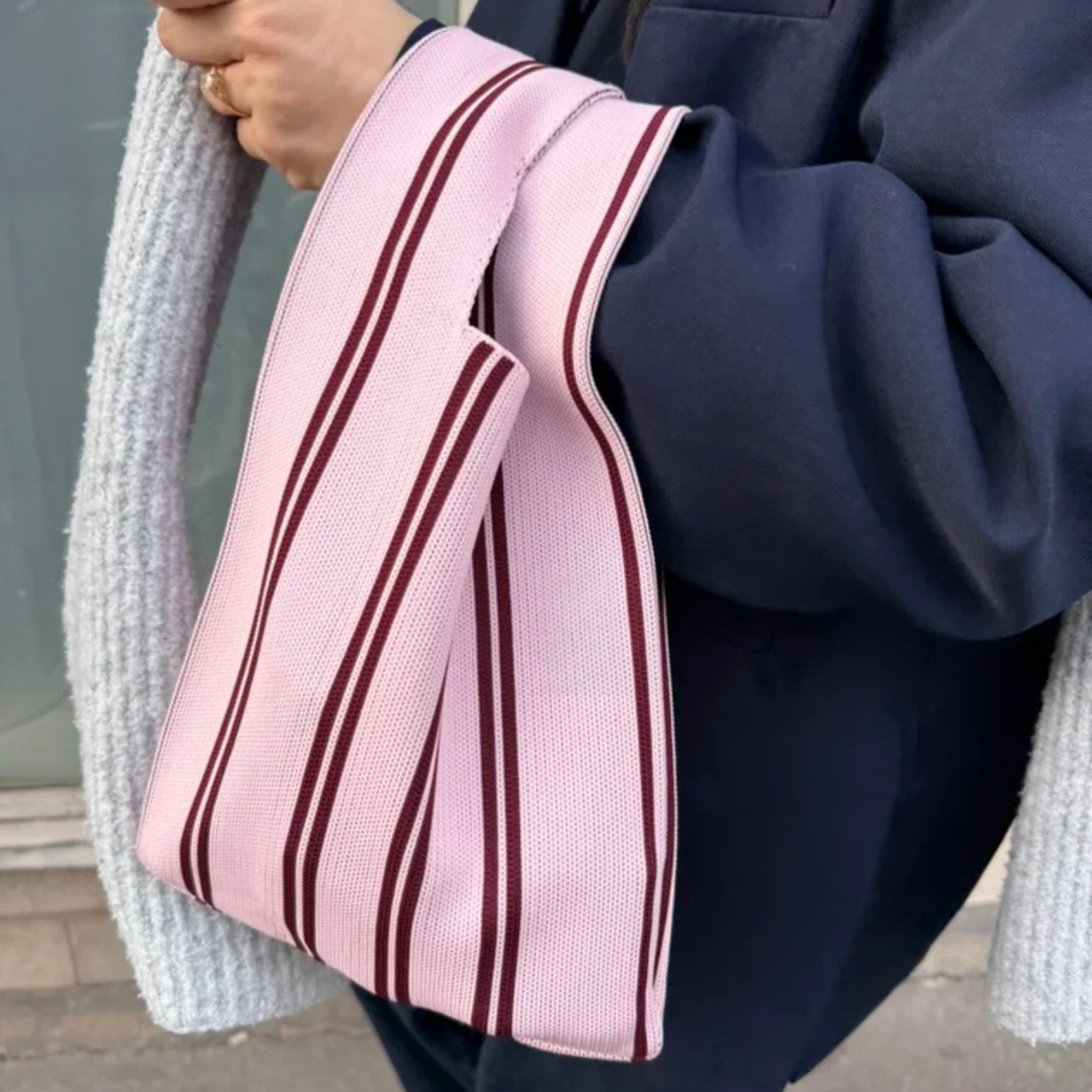 Een persoon in een donkere jas houdt een Caro Antwerp Caro Handtas Double Dash 2.0 Roland Larose vast - een roze tas met kastanjebruine strepen en grijze handvatten met structuur. Alleen de hand, een deel van de arm en de romp zijn zichtbaar.