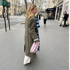Een vrouw in een lange groene jas, witte broek en beige schoenen loopt over een stadspad met de Caro Handtas Double Dash 2.0 Roland Larose van Caro Antwerp in de hand en draagt een blauw-wit gestreepte sjaal. Gebouwen en voetgangers zijn zichtbaar op de achtergrond.