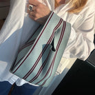 Een persoon in een wit shirt en blauwe jeans houdt de Caro Antwerp Caro Handtas Double Dash Berry Fresh vast, een lichtblauwe tas met rode en zwarte strepen en een breed handvat. Zonlicht werpt zachte schaduwen over het tafereel.