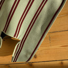 Een close-up van de Caro Handtas Double Dash Berry Fresh van Caro Antwerp. Het rood met beige gestreepte vlechtwerk rust op een houten oppervlak met duidelijke knopen en nerfpatronen.