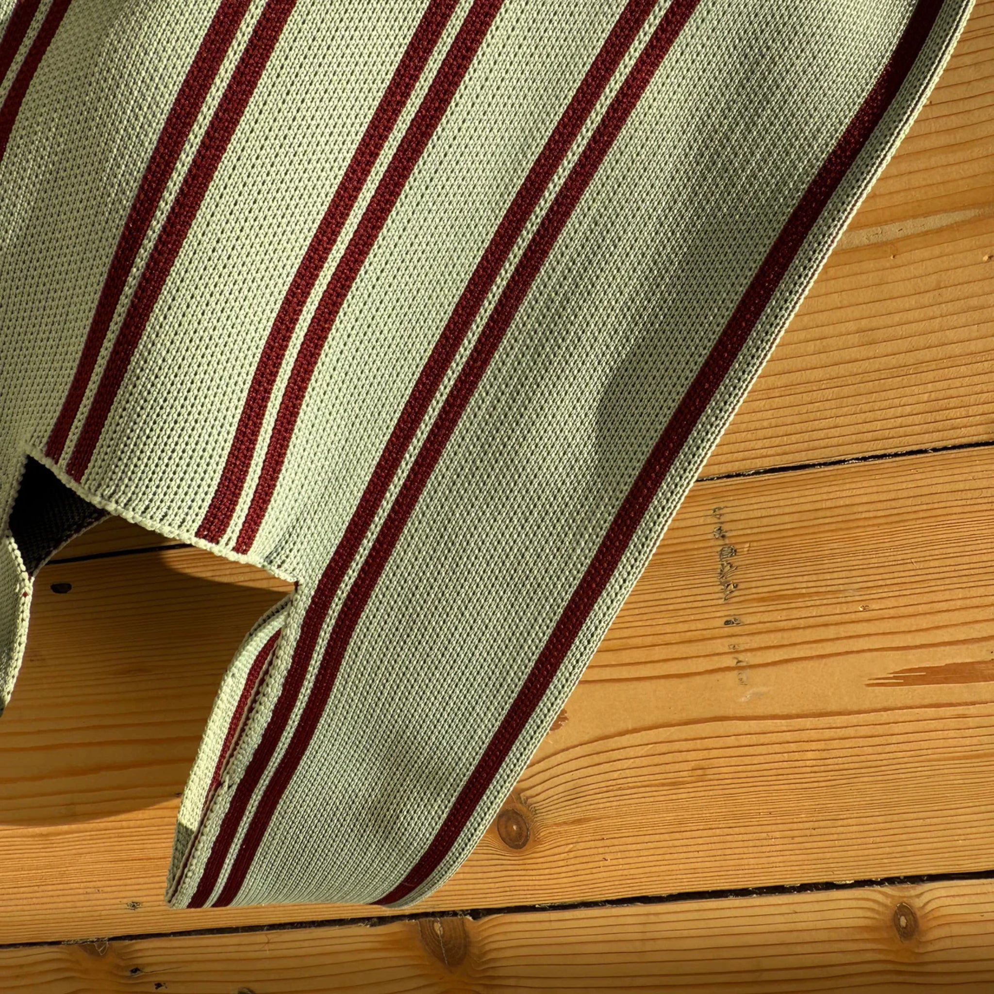 Een close-up van de Caro Handtas Double Dash Berry Fresh van Caro Antwerp. Het rood met beige gestreepte vlechtwerk rust op een houten oppervlak met duidelijke knopen en nerfpatronen.