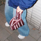 Een persoon in een blauwe spijkerbroek, een blauw jack en witte sneakers draagt de Caro Antwerp Caro Handtas Double Dash Pink Promise terwijl hij over een geplaveid trottoir naast een witte bakstenen muur loopt.