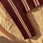 De Caro Antwerp Handtas Double Dash Pink Promise, met kastanjebruine stof met twee witte strepen, ligt op een verfrommeld beige doek en baadt in warm zonlicht.