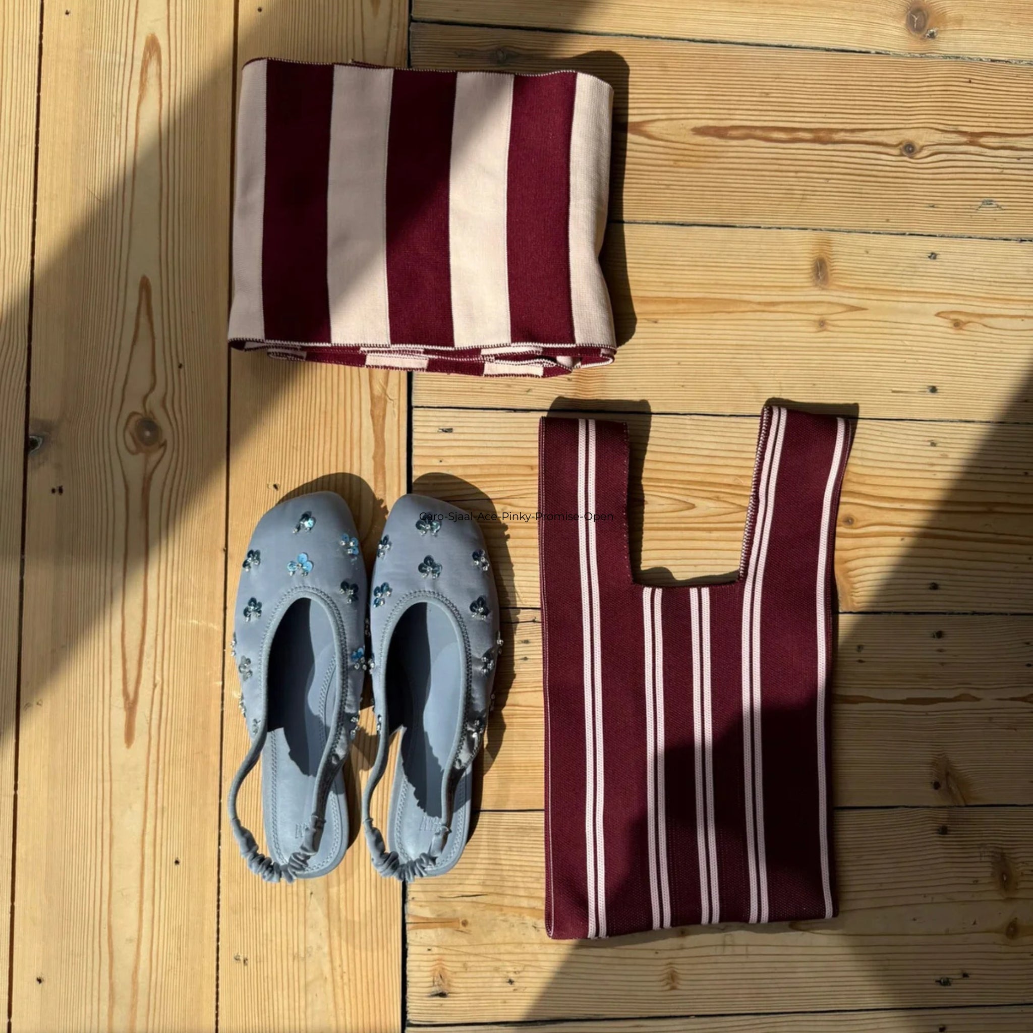 Een paar lichtblauwe versierde schoenen, een opgevouwen kastanjebruin en beige gestreepte doek en een Caro Antwerp Caro Handtas Double Dash Pink Promise staan op een houten vloer in het zonlicht.