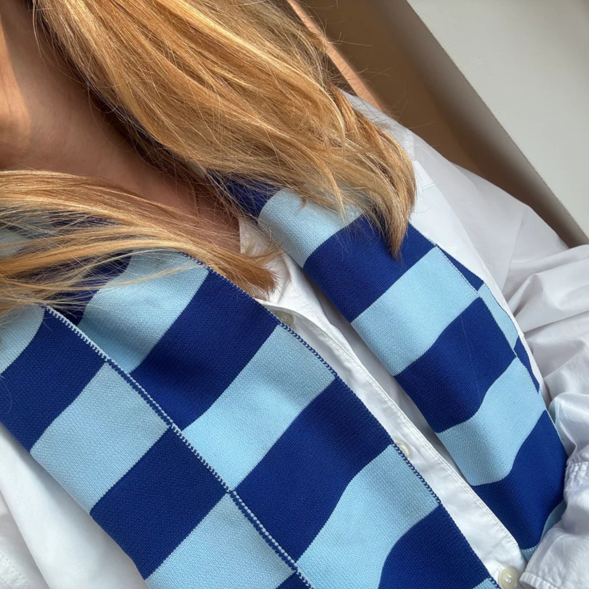 Een persoon met lang blond haar, in een wit shirt en een Caro Sjaal Ace Ice Ice Baby van Caro Antwerp, vanaf de schouders gezien.
