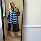 Een persoon neemt een selfie voor een spiegel en draagt een lange bruine jas, een losse witte broek, sneakers en de Caro Antwerp Caro Sjaal Ace Ice Ice Baby sjaal. De kamer heeft lichte muren en houten vloeren.