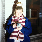 Een persoon met blond haar in een paardenstaart, een blauwe jas aan en de Caro Antwerp Caro Sjaal Ace Pinky Promise, staat tegen een witte bakstenen muur en kijkt naar zijn smartphone terwijl fel licht zijn schaduw werpt.