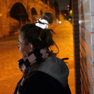 Een persoon met lichtbruin haar in een knot, draagt een Caro Antwerp Caro The Scrunch Club Scrunchie 2 st., staat 's nachts bij een betegelde muur in een schemerige straat, gekleed in een sjaal en jas.