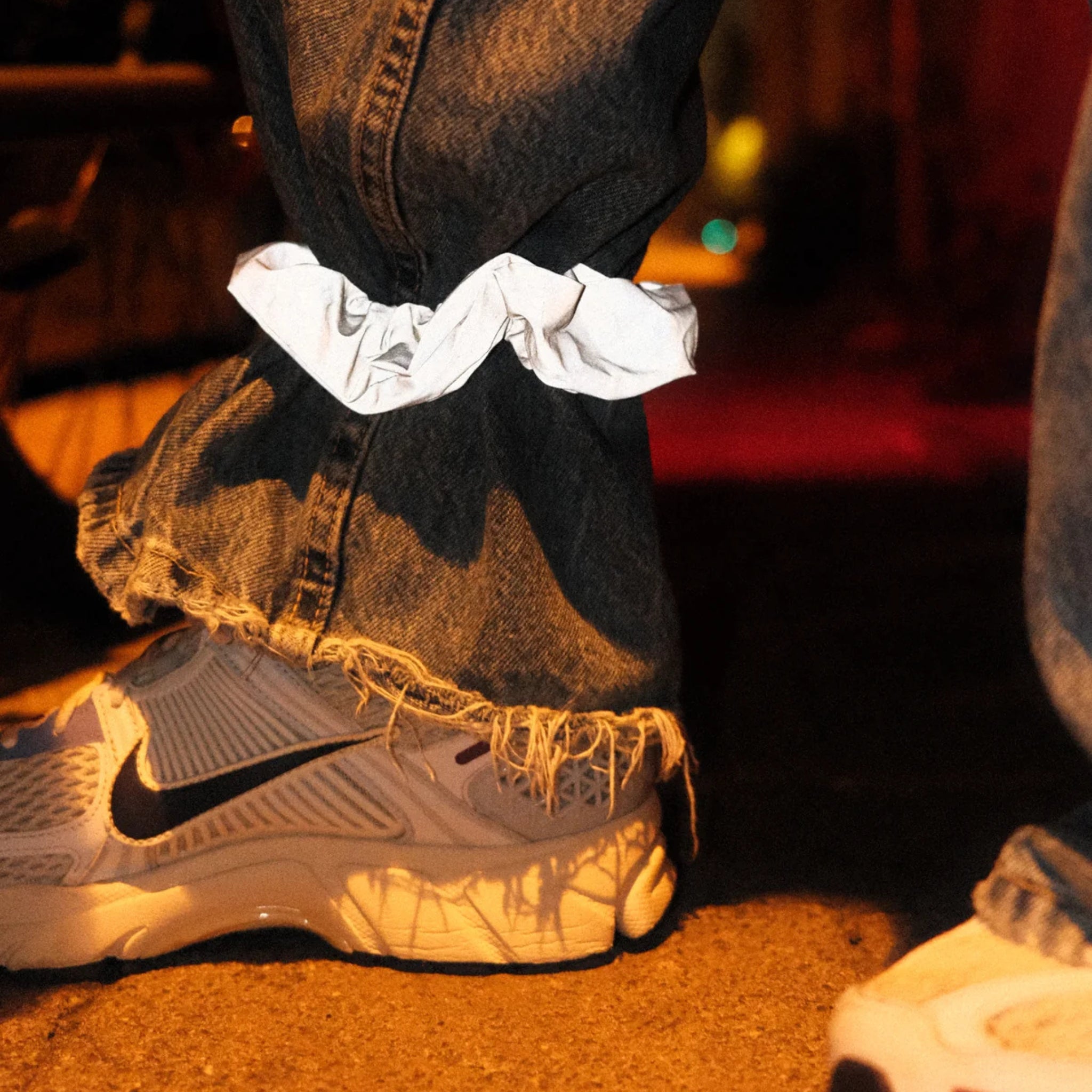 Een persoon in een gerafelde blauwe spijkerbroek en witte Nike sneakers draagt een Caro Antwerp Caro The Scrunch Club Scrunchie 2 st. om één broekspijp. De schemerige scène heeft een warme, stemmige sfeer.