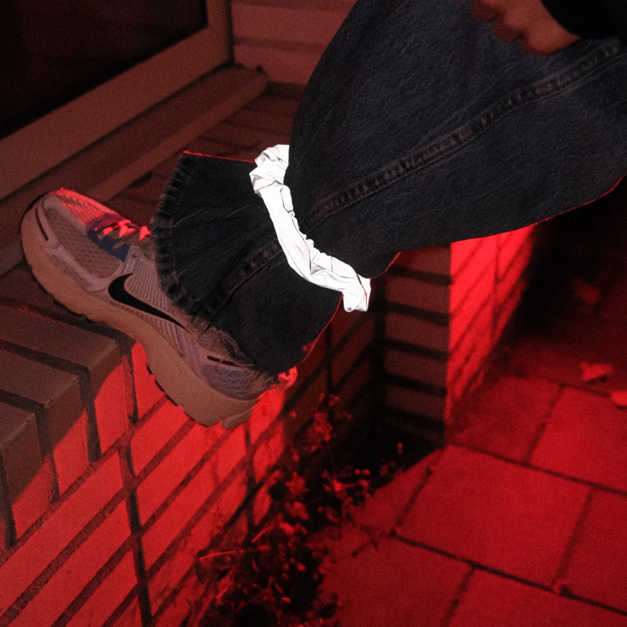 Een persoon in jeans en witte sneakers stapt 's nachts op een stenen richel. Hun spijkerbroek is vastgemaakt met een Caro Antwerp Scrunch Club Scrunchie, de perfecte fietsaccessoire, terwijl rode verlichting de scène verlicht.