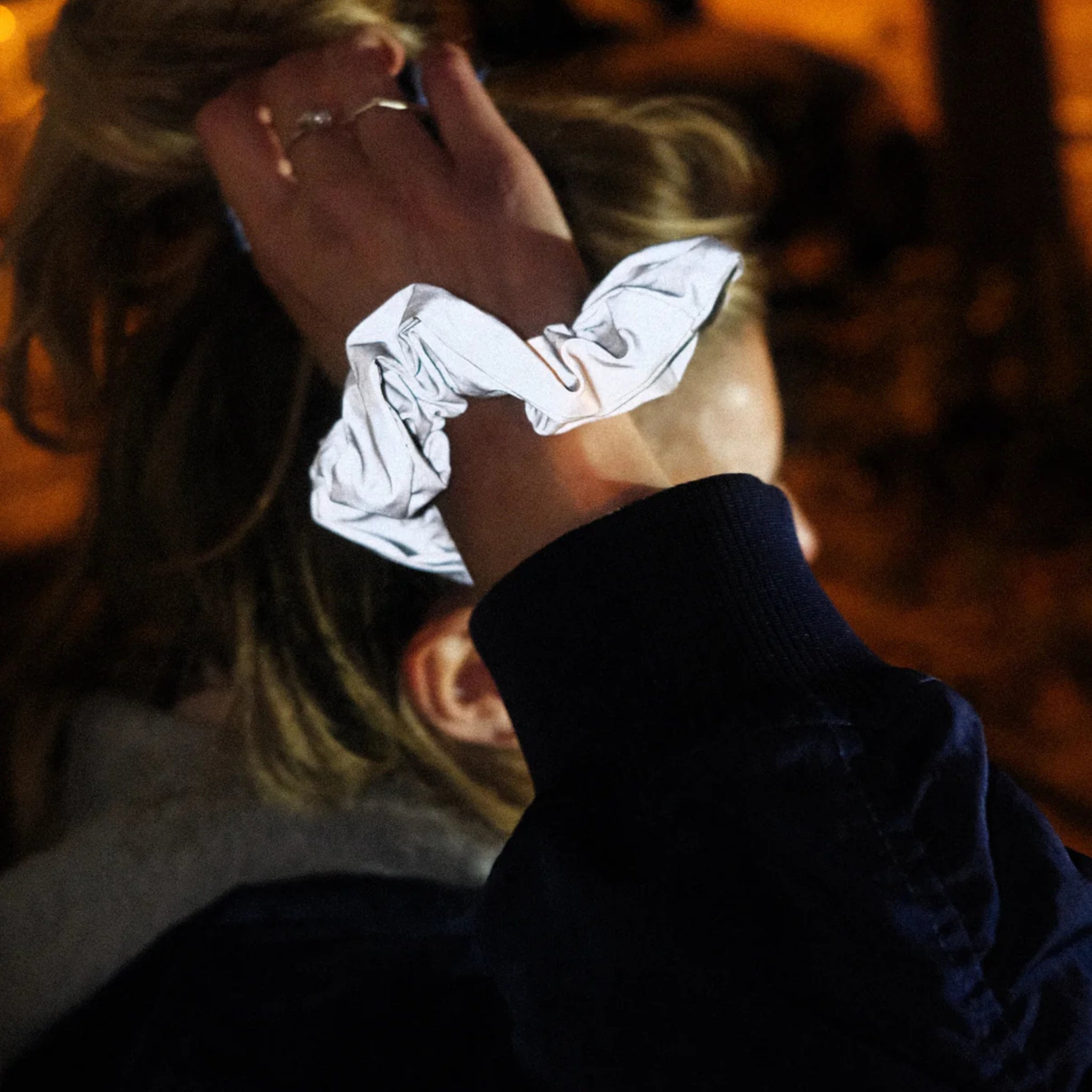 Een persoon met blond haar, gedeeltelijk verborgen, showt de reflecterende Caro Antwerp Caro The Scrunch Club Scrunchie 2 st. - een perfect fiet accessoire - tegen een warm verlichte, onscherpe achtergrond.