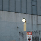 De Fine Lumens Mono Leeslamp LED Oplaadbaar in olijfgroen-geel van Fine Lumens staat op een beige voet naast een betonnen muur met waarschuwingsborden, voor een gebouw met een metalen gevel in rasterpatroon.