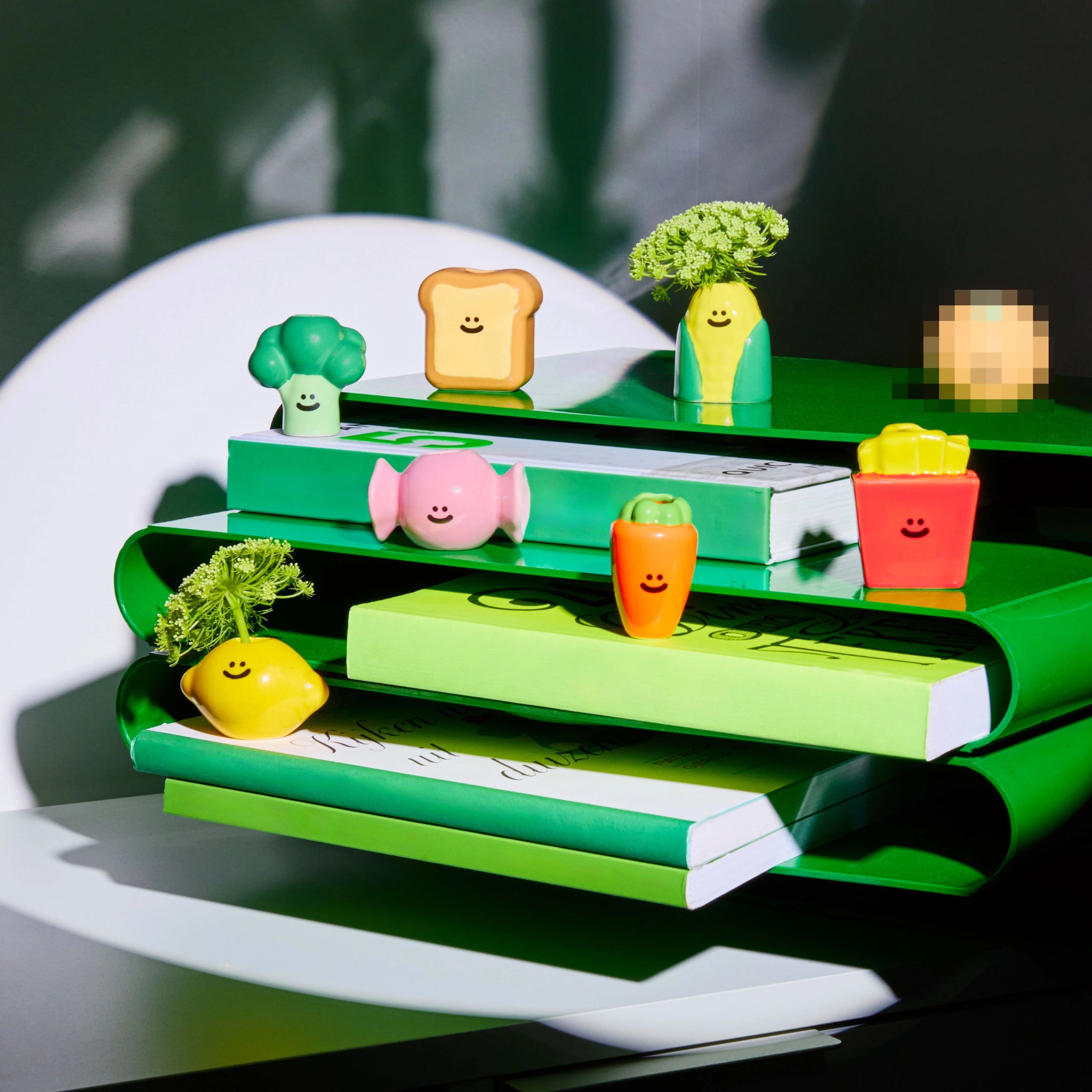 Een groep kleurrijke, lachende speelgoedgroenten en -voedsel, waaronder broccoli, maïs, toast, frietjes, citroen, wortel, een varken en het Fluid Market Box Food Vaasje "Verrassing" 1 st. van Fluid Market, staat op groene en witte boeken; één achtergronditem is gepixeld.