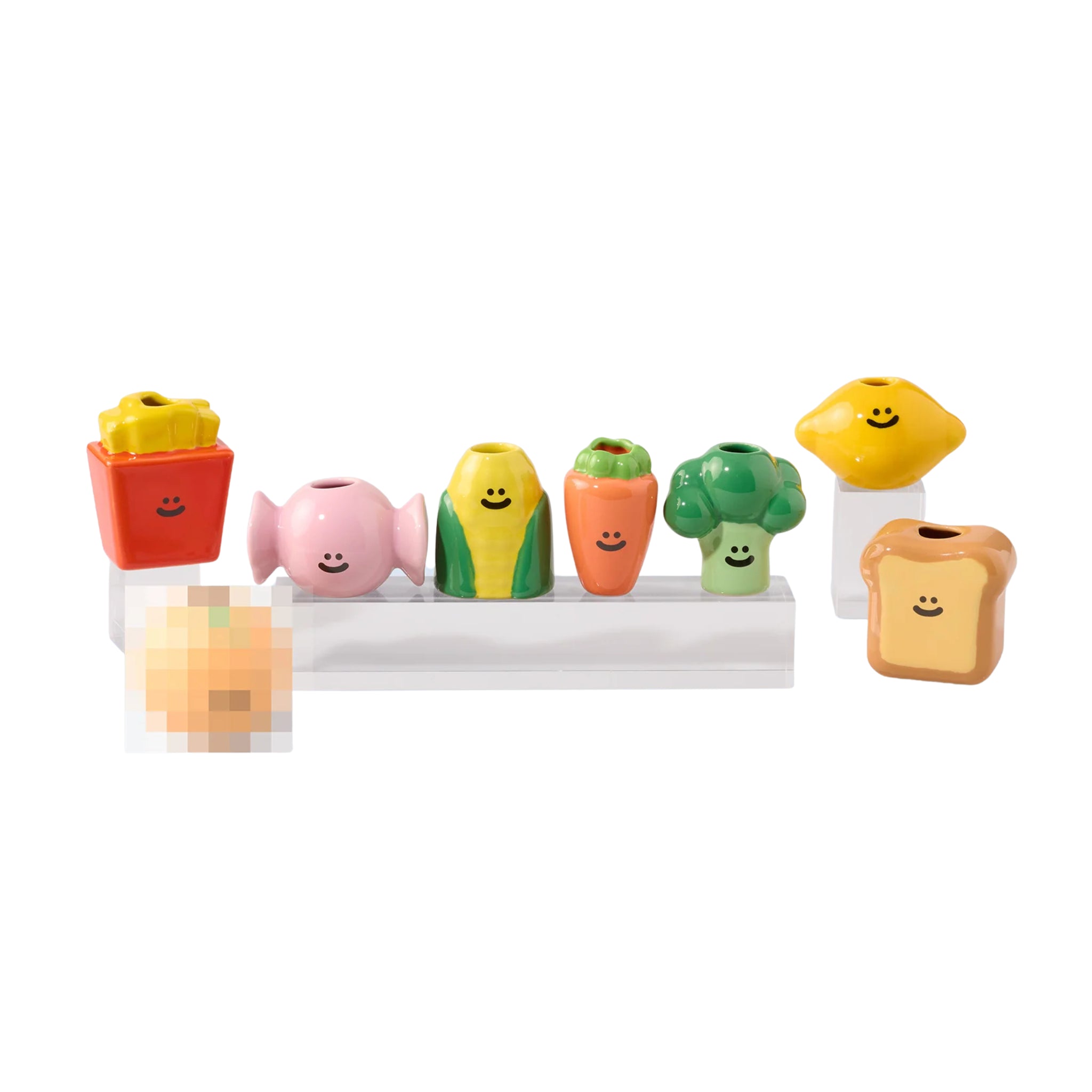 Het Fluid Market Box Food Vaasje "Verrassing" van Fluid Market heeft zeven kleurrijke, lachende objecten van keramiek in de vorm van friet, snoep, maïs, wortel, broccoli, citroen en brood op een rij met één vaag voorwerp ervoor.