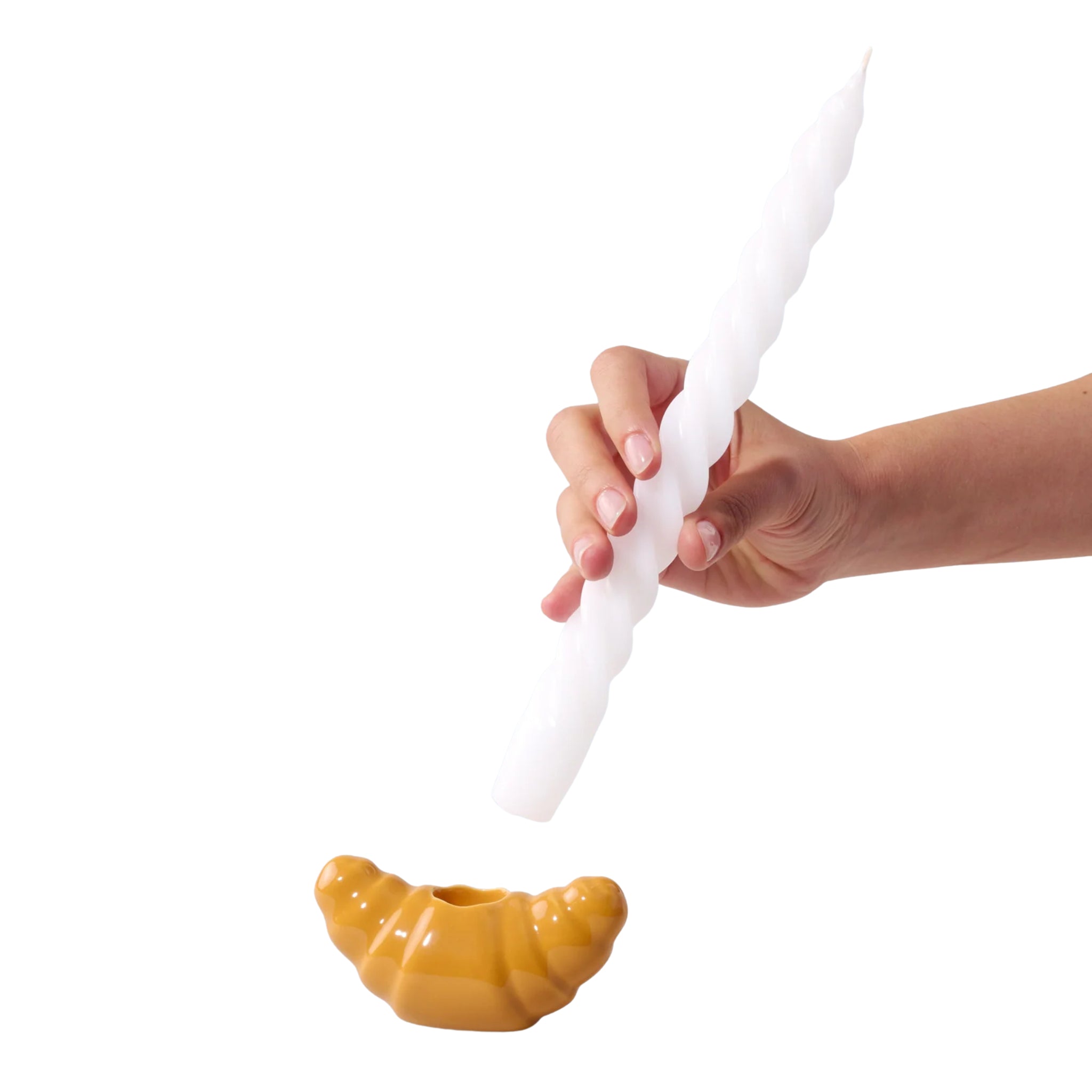 Een hand houdt een lange, gedraaide witte kaars vast boven de Fluid Market Croissant Kandelaar van Fluid Market, een unieke kandelaar van keramiek tegen een effen witte achtergrond.