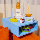 Op een blauw bijzettafeltje staat Fluid Market Zout- en Pepervaatjes Baguette 2 st. als speels tafelaccent, plus boeken, een kleine wijnfles en een straatnaambordje RUE BON APPETIT. Oranje kussens en transparante gordijnen maken het gezellige tafereel compleet.