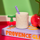 De Fluid Market Mok Kers van Fluid Market, met kersvormig handvat, bevat een paarse lepel en staat op het lavendelkleurige boek 'PROVENCE GLORY' voor een groene muur met kleurrijke objecten op de achtergrond.