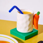 De Fluid Market Mok Wortel van Fluid Market - met een wortelvormig handvat - bevat sinaasappelsap en een blauw rietje op een groene onderzetter in een gele kleur. Een bord met een sinaasappelschijfje steekt naar voren, waardoor deze mok een origineel cadeau is voor ontbijtliefhebbers.