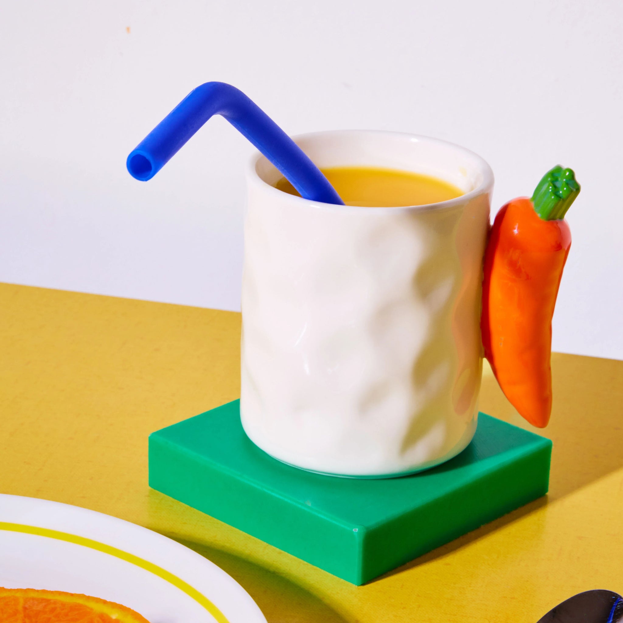 De Fluid Market Mok Wortel van Fluid Market - met een wortelvormig handvat - bevat sinaasappelsap en een blauw rietje op een groene onderzetter in een gele kleur. Een bord met een sinaasappelschijfje steekt naar voren, waardoor deze mok een origineel cadeau is voor ontbijtliefhebbers.