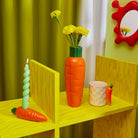 Een gele houten plank toont een wortelvormige vaas met gele bloemen, een groene spiraalkaars in een wortelhouder, de Fluid Market Mok Wortel van Fluid Market en een klein worteldecor. Aan de groene muur hangt een rode spiegel - perfect als origineel cadeau.