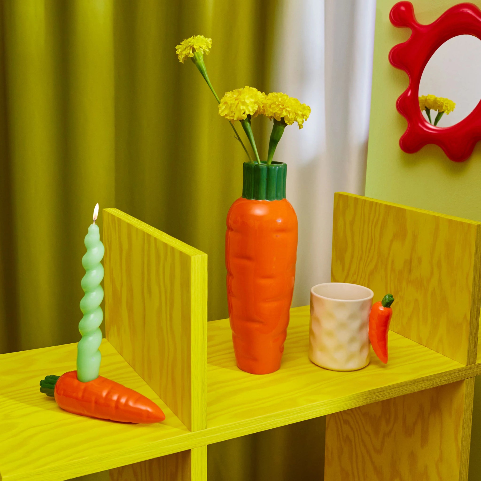 Een gele houten plank toont een wortelvormige vaas met gele bloemen, een groene spiraalkaars in een wortelhouder, de Fluid Market Mok Wortel van Fluid Market en een klein worteldecor. Aan de groene muur hangt een rode spiegel - perfect als origineel cadeau.