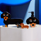 De Fluid Market Zout- en Pepervaatjes Teckel 2 st. -zwarte en bruine teckels van keramiek met cartoongezichtjes- worden weergegeven op een wit oppervlak tegen een blauwe achtergrond, wat zorgt voor een speels en leuk cadeau.