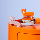 Twee keramieken katjes van Fluid Market, één oranje en één lichtgrijs, uit de Fluid Market Zout- en Pepervaatjes Kat 2 st. set staan op een oranje cilinder, perfect als design cadeau tegen een lichtblauwe achtergrond.