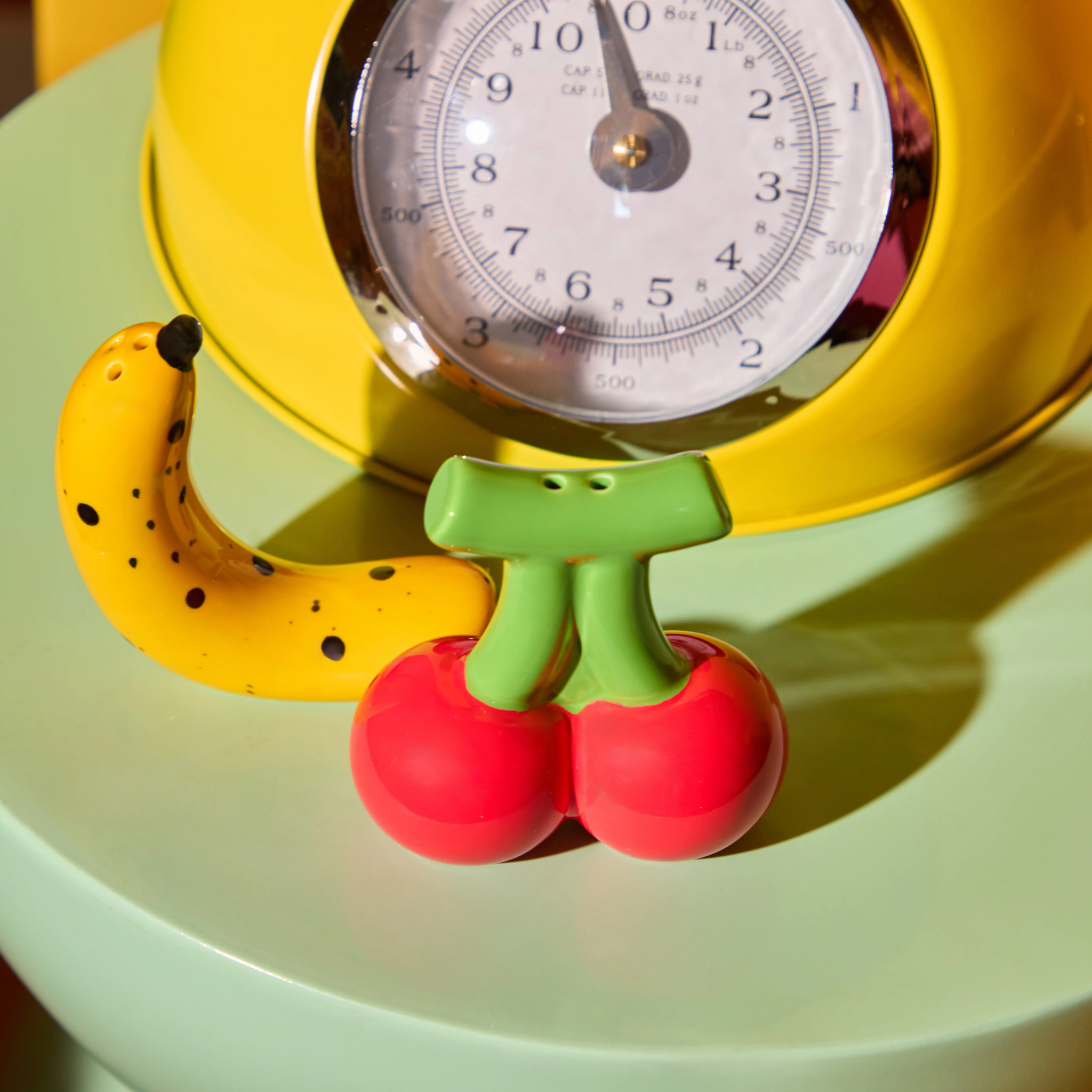 De Fluid Market Zout- en Pepervaatjes Banaan Kers 2 st. set rust op mintgroen, omlijst door een retro gele keukenschaal - perfect om speelse charme toe te voegen aan de keukeninrichting van elke Fluid Market fan.
