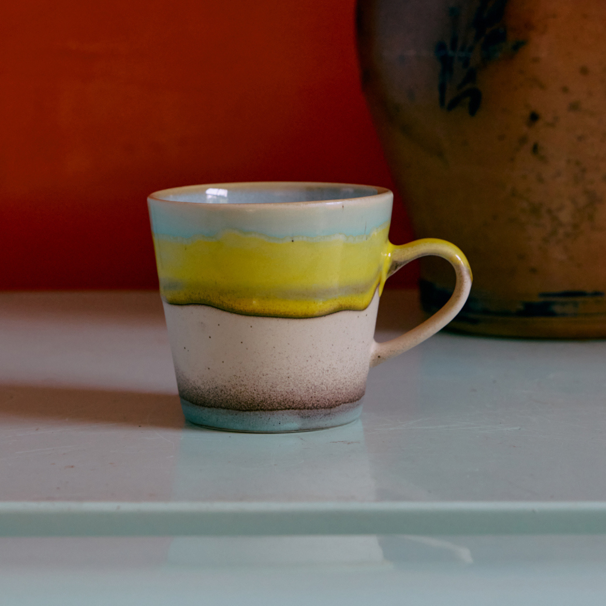 De HK Living 70s Ceramics Americano Mok Glint, met blauwe, gele en bruine golvende strepen, staat op een lichtblauwe ondergrond, met op de achtergrond een grote bruine vaas tegen een oranje muur.