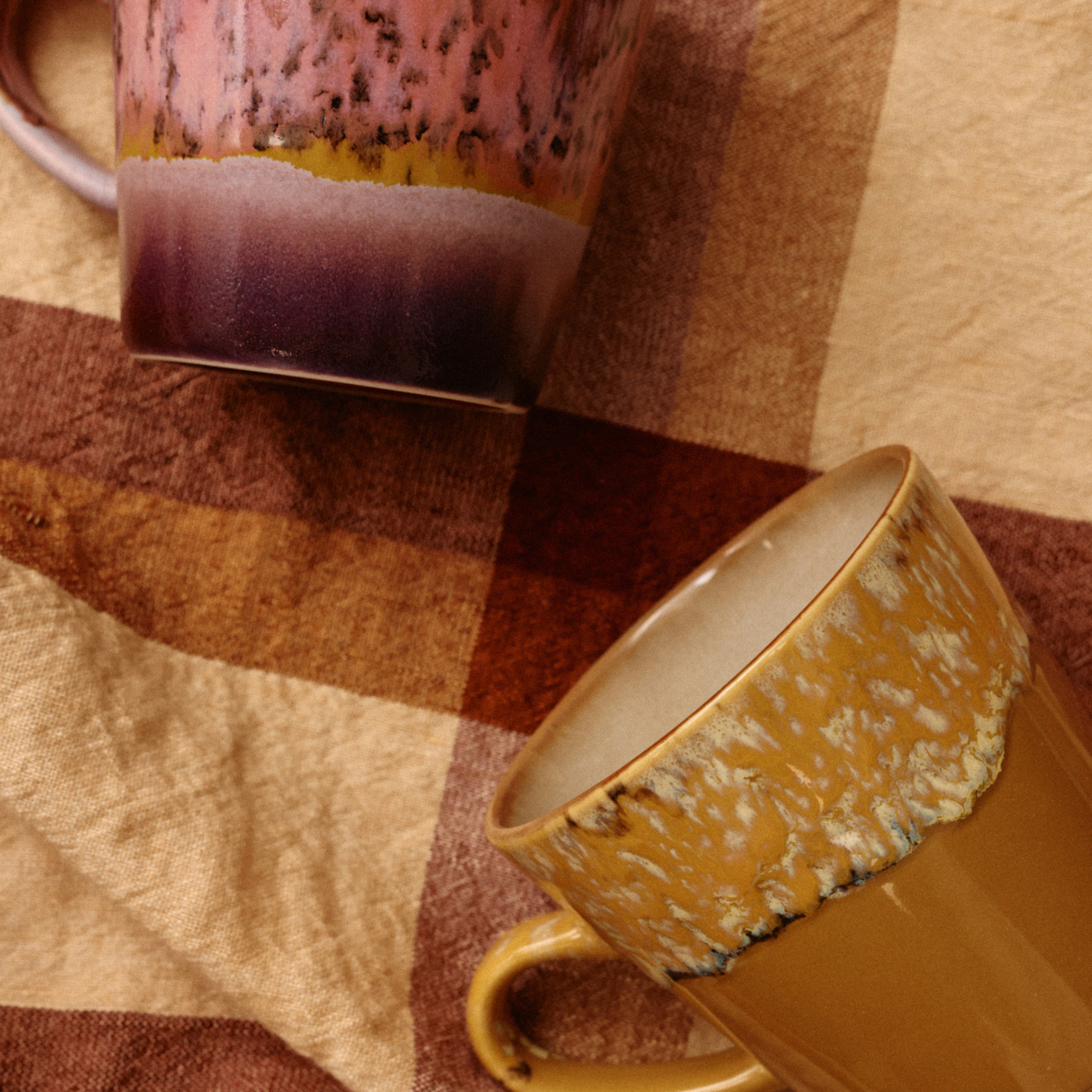De HK Living 70s Ceramics Americano Mok Sunbeam is voorzien van gevlekte, aardse glazuren in roze, paars, geel en bruin, afgezet tegen een bruin-beige geruite stof — wat de charme van een retro koffiemok oproept.