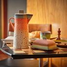 Een HK Living 70s Ceramics Botervlootje Mercury botervlootje staat met een keramische kan, beker, brood en servetten op een geruite tafel. Een gezellige, warm verlichte sfeer met een houten stoel en muur op de achtergrond.