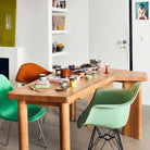 Een houten eettafel met levendig keramiek, waaronder het HK Living 70s Ceramics Botervlootje Mercury, wordt omringd door een oranje en twee groene stoelen in een moderne kamer met boekenplanken en kunst aan de muren.