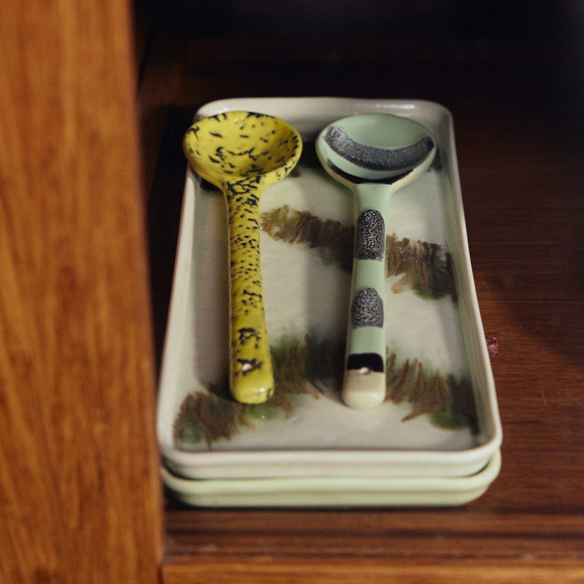 Twee 70s Ceramics lepels - een gele met zwarte spikkels, een groene met donkere accenten - liggen op een HK Living 70s Ceramics Cake Schaal Tide met groene en bruine motieven, die samen met twee andere op een houten plank staat.