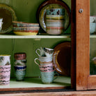 Een lichtgroene houten kast met een glazen deur toont de HK Living 70s Ceramics Cappuccinokop Dada 4 st. set, kommen en borden - allemaal met aardgroene, roze, beige en blauwe glazuren van HK Living.