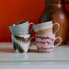 Een set HK Living 70s Ceramics Cappuccinokop Dada 4 st. mokken met unieke, kleurrijke glazuurpatronen zijn gestapeld op een wit oppervlak, met een aardebruine kan en een oranje muur op de achtergrond.