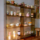 Glazen planken tonen netjes HK Living 70s Ceramics Koffiekop Bubble en andere handgemaakte keramische stukken. Zonlicht accentueert het kleurrijke aardewerk, met ingelijste kunst aan de muur naast de planken.