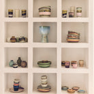 Een witte ingebouwde plank met twaalf vierkante vakjes toont kleurrijke HK Living 70s Ceramics Koffiekop Bramble - elk met de hand gemaakt met unieke texturen, netjes gerangschikt in elk vakje.