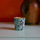 De HK Living 70s Ceramics Koffiekop Patina, een blauw met bruin gespikkelde beker van HK Living, staat op een licht oppervlak met een grote bruine vaas erachter tegen een oranje muur.