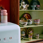 Een pastelkleurige keuken toont een retro Smeg koelkast met daarop een keramische pot en een open kast met een assortiment keramiek in aarde- en pasteltinten, waaronder de HK Living 70s Ceramics Noodle Kom Lush D13 kom.