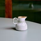 Het HK Living 70s Ceramics Melkkannetje Petal, een klein wit kannetje van keramiek met een handvat, staat op een gladde lichtgrijze tafel. De zacht vervaagde groene en bruine achtergrond benadrukt de charme van dit handgemaakte servies.