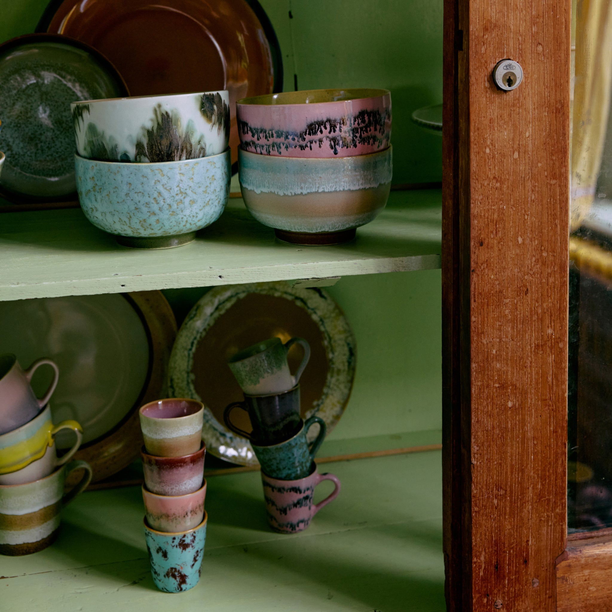In een houten kast met glazen deuren staan HK Living 70s Ceramics Noodle Kom Bubble D13 kommen, gestapeld met borden in aardetinten en getextureerde glazuren, allemaal netjes gerangschikt op lichtgroene planken.