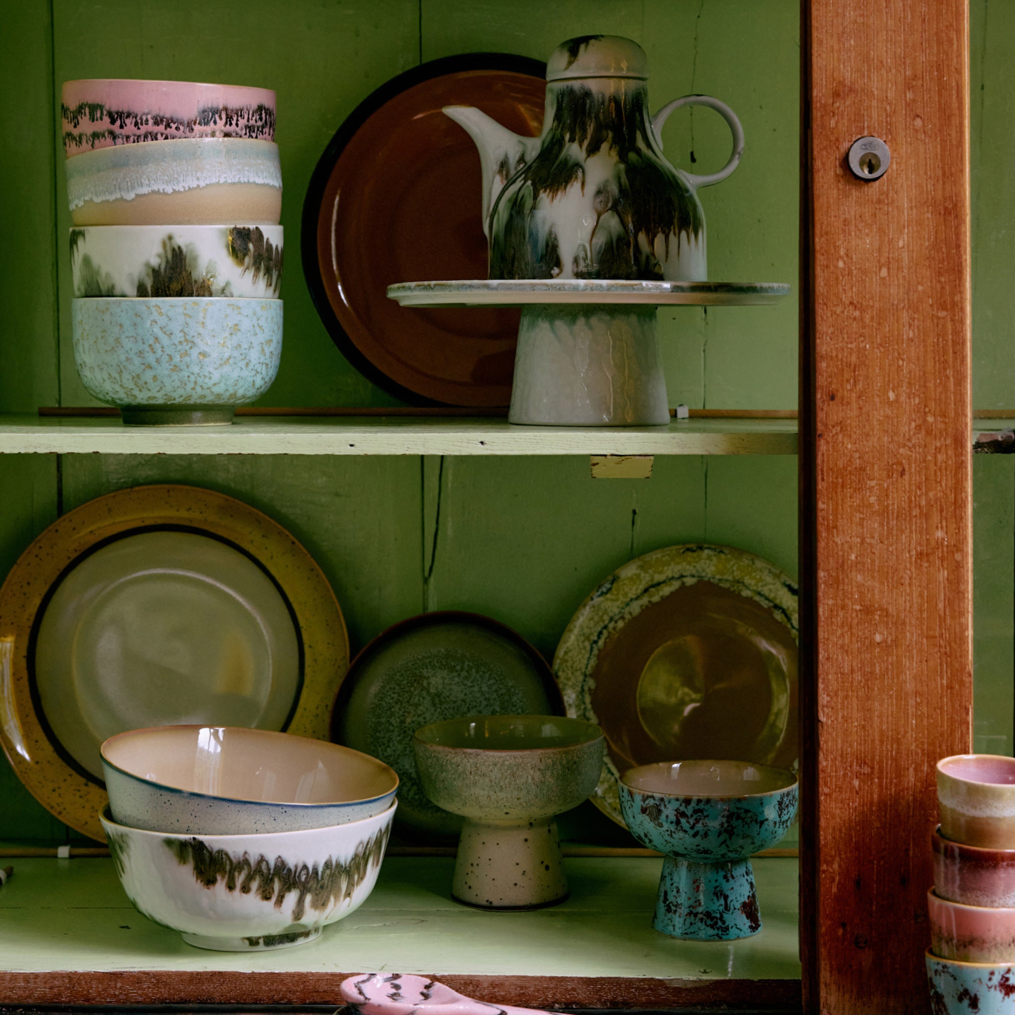 In een houten kast met groene planken staan HK Living 70s Ceramics Noodle Kom Dada D13 4 st. -distinctieve keramische kommen in aardetinten, elk met unieke glazuurpatronen.