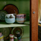 In een houten kast met lichtgroene planken staan HK Living 70s Ceramics Noodle Kom Dada kommen (set van 4) in levendige patronen. De iets geopende deur onthult een slot aan de rechterkant.