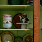 De HK Living 70s Ceramics Plateau/Taartschaal Smudge M heeft retro groene, roze en bruine patronen en voegt vintage stijl toe aan lichtgroene planken in een houten kast met glazen deur.