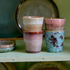 Vier HK Living 70s Ceramics Ristretto Mok Futurism kopjes staan paarsgewijs op een lichtgroene plank, met borden en een ondiepe kom erachter. Elk kopje heeft kleurrijke, gespikkelde glazuren in aardetinten van HK Living.