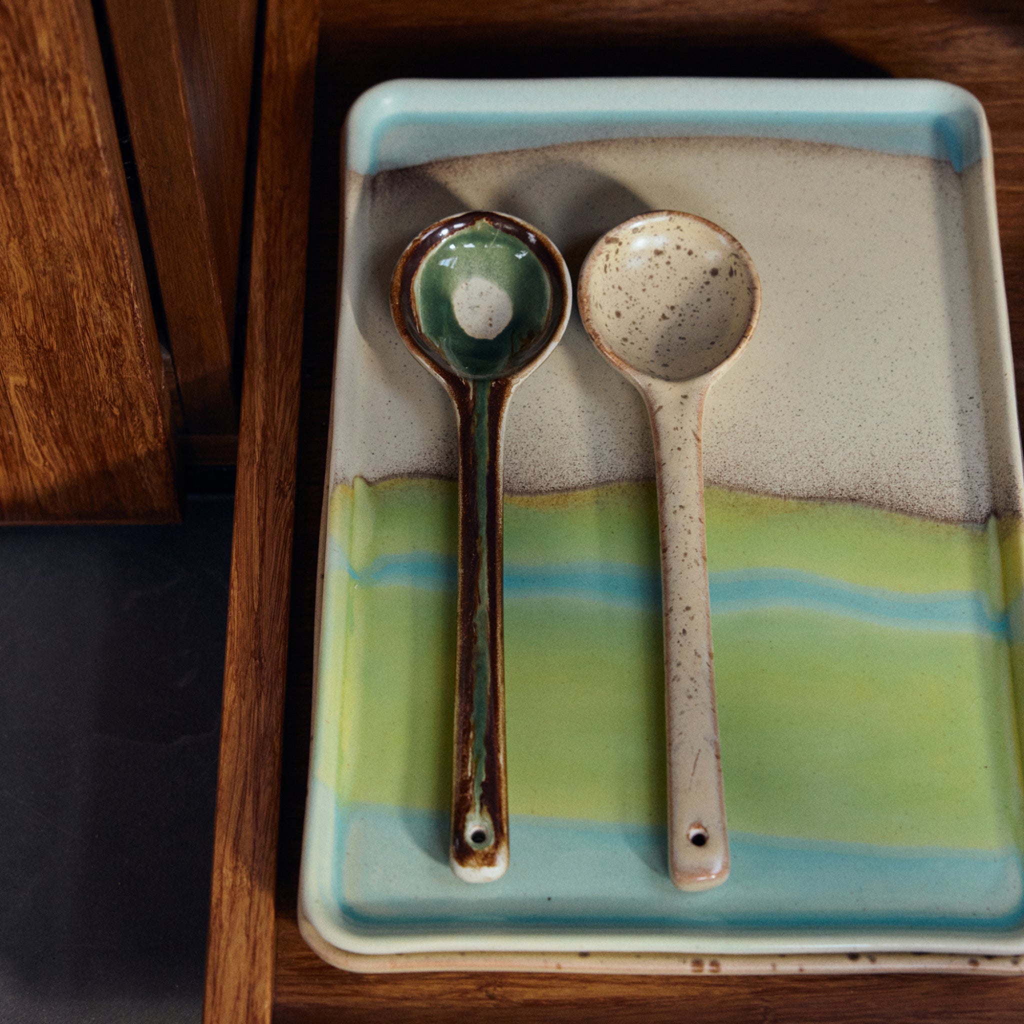 Twee keramische soeplepels met verschillende glazuurontwerpen rusten naast elkaar op een HK Living 70s Ceramics Schaal Glint L, een rechthoekige, veelkleurige schaal met groene, blauwe en bruine tinten.