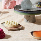 Een modern stilleven met HK Living 70s Ceramics Schaaltje Atlas (2 stuks), een gespikkelde taartstandaard, glitterbol, bellenobject op een schaal en andere levendige retro-geïnspireerde voorwerpen op een licht geweven tafelkleed.